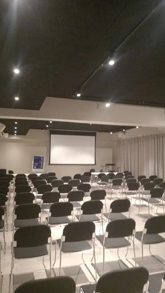Conference room with rows of dark chairs facing a projection screen under a black ceiling with spotlights