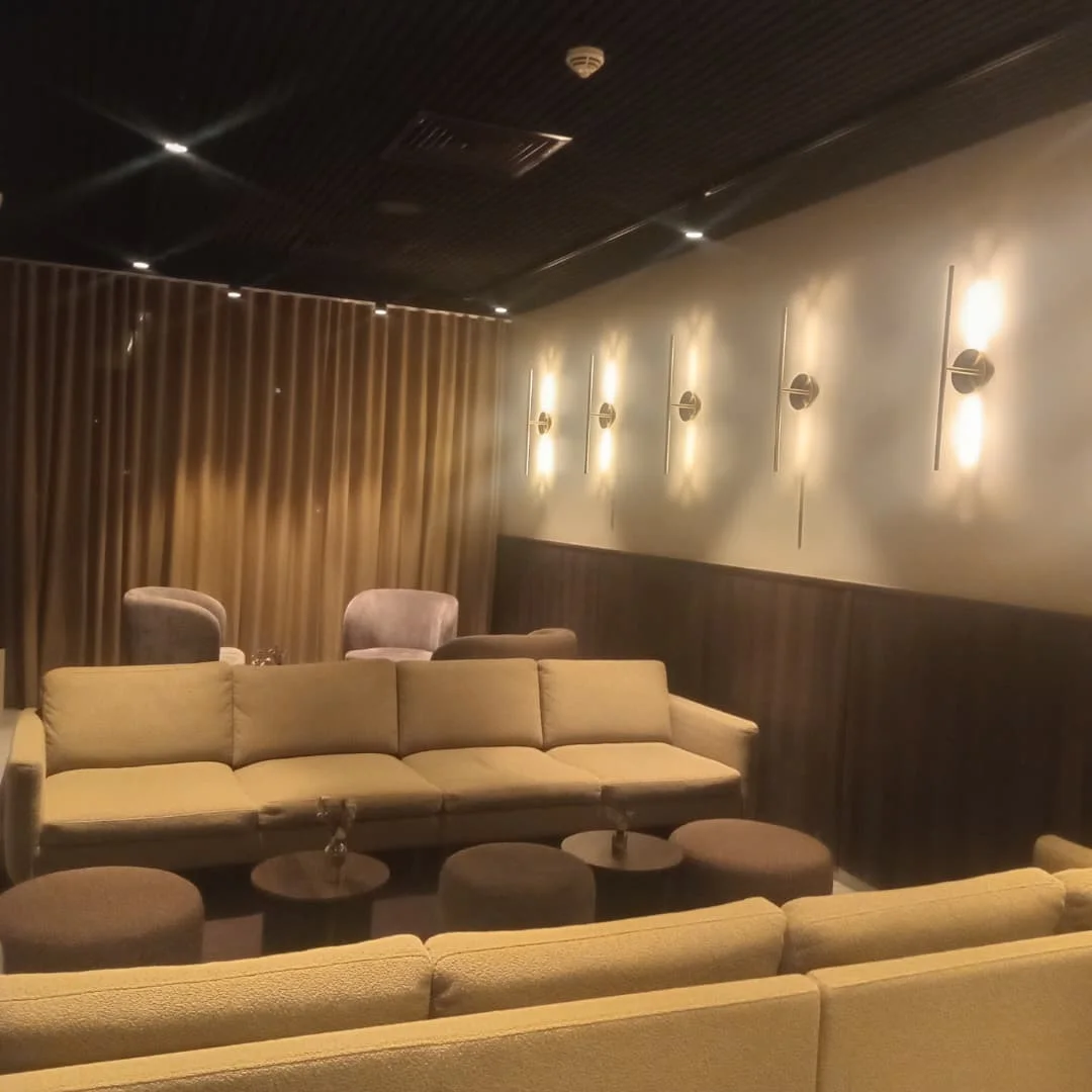 Hotel lounge with a beige sofa, round stools and wall sconces beneath a dark ceiling with recessed spotlights